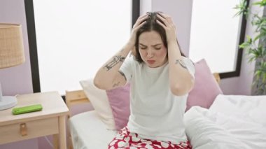 Pijama giymiş stresli genç bir kadın yatağa oturup ağrılı migren ağrısı çekiyor. Eller başının üstünde, şiddetli baş ağrısı stresi altında, yatak odasında acı çekiyor..