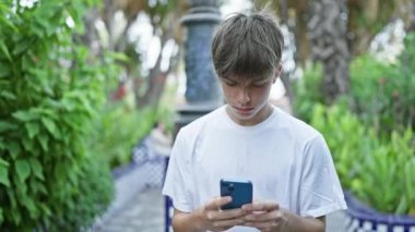 Beyaz tişörtlü genç, yemyeşil bahçe yolunda akıllı telefon kullanıyor.