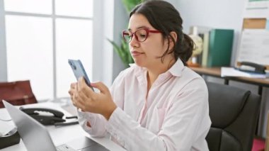 Ofis ortamında genç bir İspanyol kadın, modern iş yerinde akıllı telefonuna dikkatlice bakıyor..