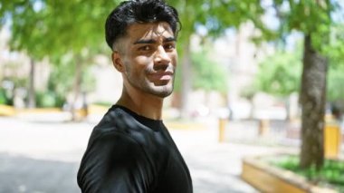 Sakallı yakışıklı bir genç, doğal bir karizma yayarak, yemyeşil bir parkta gelişigüzel gülümsüyor..