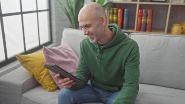 Yeşil kazaklı kel adam tablet kullanıyor ve evdeki gri kanepede kahve içiyor.