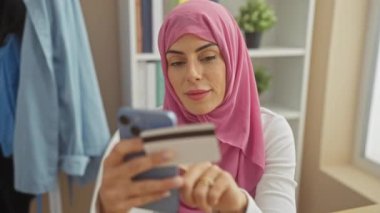 Evde kredi kartı ve akıllı telefon kullanan pembe tesettürlü bir kadın rahat bir iç mekanda e-ticaret ve teknoloji sergiliyor..