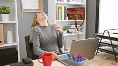 Gözlüklü, gülen sarışın kadın ofis masasında dizüstü bilgisayar, kapalı bitkiler ve kırmızı bardakla eğleniyor.