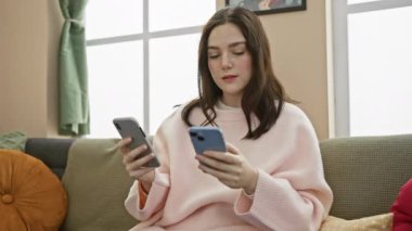 Pembe kazaklı genç bir kadın evdeki kanepede rahatça akıllı telefonlar kullanıyor..
