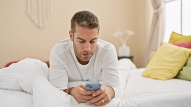 Yatak odasında akıllı telefon kullanan, etkileyici yüzü ve beyaz yatağı olan bir adam.