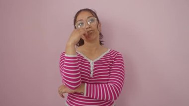 Düşünceli, genç İspanyol bir kadın çizgili tişört giyiyor, eli çenesinde, düşünceli tefekküre dalmış, kafa karıştırıcı bir fikri sorguluyor, izole edilmiş pembe arka plana karşı.