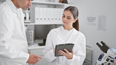 Bilim adamı olarak tasvir edilen bir erkek ve kadın modern bir laboratuvar ortamında bir tablet kullanarak işbirliği yapıyorlar..