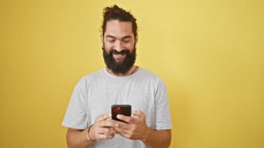 Sarı duvara karşı akıllı telefon kullanan yakışıklı sakallı adam, neşe ve onay gösteriyor..