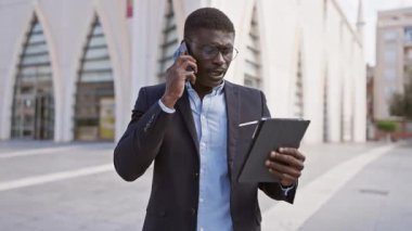 Takım elbiseli profesyonel bir zenci akıllı telefon kullanır ve şehir caddesinde bir tablet bulundurur..