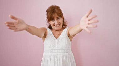 Pembe elbiseli neşeli orta yaşlı kadın, sıcak bir kucaklama için kolları açık! Mutlulukla ışıldıyor, pembe bir duvara yaslanıp seni kucaklamak için içeri davet ediyor.!