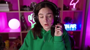 Yeşil kapüşonlu genç bir kadın, geceleri neon ışıklı bir oyun odasında mikrofonlu bir kulaklık kullanıyor. Sıradan bir oyuncu ya da flamayı canlandırıyor..