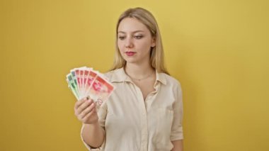 Heyecanlı genç sarışın kadın, fikirlerle ve sevinçle dolup taşıyor, şaşırtıcı bir şekilde İsrail şekeli banknotlarını gösteriyor. Mutlu, düşünen, sarı bir arka planda izole edilmiş bir yüz..