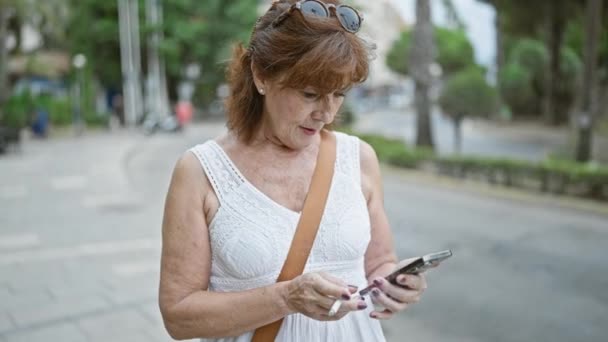 在城市街道上使用智能手机的红头发女士 戴着太阳镜 身穿白色衣服 — 图库视频影像