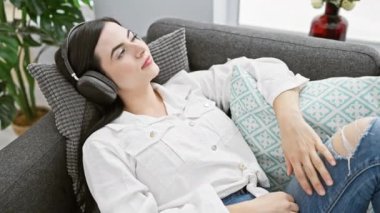 Çekici genç İspanyol kadın, modern bir oturma odasında gri koltukta, kulaklıklarıyla dinleniyor..