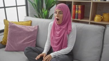 Şaşırmış genç İspanyol kadın inanılmaz bir şaşkınlık yaşıyor, şok ve korku dolu duygularla yüzleşiyor, tesettür giyiyor, ağzı açık bir şekilde kanepede oturuyor.