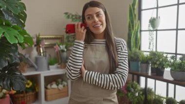 Gülümseyen bir kadın, bitkilerle dolu bir iç odada, sıcak ve rahat bir atmosfer yayarak telefonla konuşmaktan zevk alır..