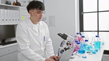 Laboratuvar önlüğü giymiş kendine güvenen genç bir adam mikroskop ve beherlerle modern bir laboratuvarda dizüstü bilgisayarda çalışıyor..