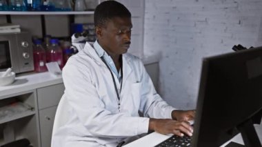 Laboratuvar önlüğü giymiş Afrikalı bir adam modern bir laboratuvar ortamında bilgisayarın başında çalışıyor..