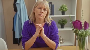 Endişeli orta yaşlı, mor kazaklı bir kadın evde oturmuş düşünceli bir ifadeyle ve arka planda ev bitkileriyle..