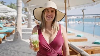 Güneş şapkalı gülümseyen kadın lüks havuz kenarındaki plaj barında mojito yudumluyor.