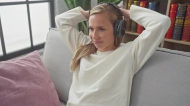 Rahat bir kadın kulaklıkla müzik dinliyor, rahat bir oturma odasındaki koltukta uzanıyor.