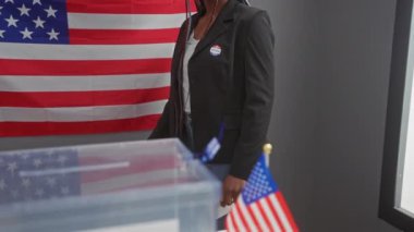 Örgü örgülü Afrikalı Amerikalı kadın Amerikan oy kullanma merkezinde oy veriyor, vatanseverlik belirgin bir şekilde gösterilmiş bayrağıyla.