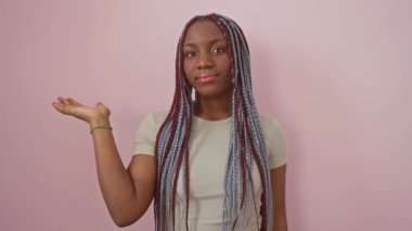 Gülümseyen, neşeli Afrikalı Amerikalı kadın avucuyla bir şey sunuyor, izole edilmiş pembe bir arka planda durup, davetkar bir şekilde kameraya bakıyor..