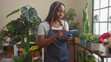Örgülü odaklanmış siyah bir kadın yeşil bitkiler ve renkli çiçeklerle dolu bir çiçekçinin içinde tablet kullanıyor..