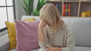 İçerideki koltukta oturan stresli bir kadın, parlak bir oturma odasında endişe belirtileri gösteriyor..