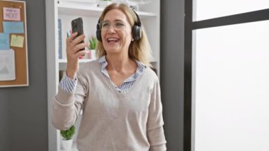Modern bir ofiste elinde akıllı bir telefonla müzik dinleyen, kulaklıklı, gülümseyen olgun bir kadın.