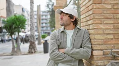 Sıradan, sakallı, kep takan rahat yetişkin bir adam, şehir ortamında dışarıda dikilirken rahatça gülümsüyor..