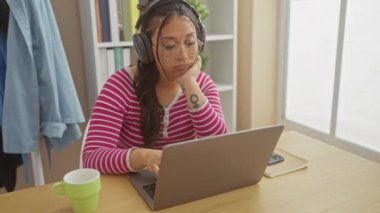 Gözlüklü ve kulaklıklı sıkılmış Latin bir kadın elinde yeşil kupa ve akıllı telefonuyla evde oturuyor..