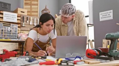 Bir erkek ve kadın, ahşap işçiliği aletleriyle çevrili bir atölyede bir dizüstü bilgisayar projesine odaklanarak iş birliği yapıyorlar..