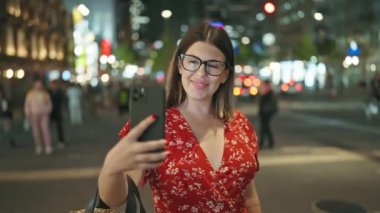 Neşeli ve güzel İspanyol bir kadın, neşeli bir anda yakalandı, akıllı telefonuyla şehir osaka sokağının gece ışıkları altında mesaj atıyor, bağlı bir dijital dünyanın özünü somutlaştırıyor..