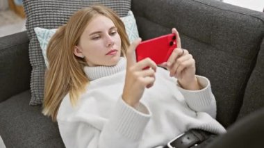 Sarı saçlı ve mavi gözlü genç bir kadın evdeki akıllı telefonunu kullanarak koltukta dinleniyor..