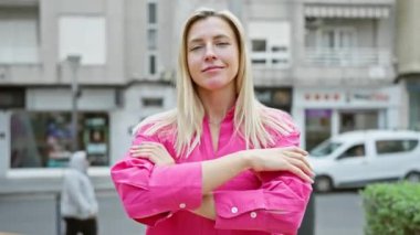 Kendine güvenen, sarı saçlı, parlak pembe tişörtlü beyaz bir kadın şehir sokaklarında kollarını kavuştururken gülümsüyor..