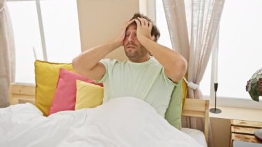 Yatak odasında şiddetli baş ağrısı çeken stresli beyaz bir adam. Eller başının üstünde, pijamada ağrı ve gerilimi ifade ediyor..