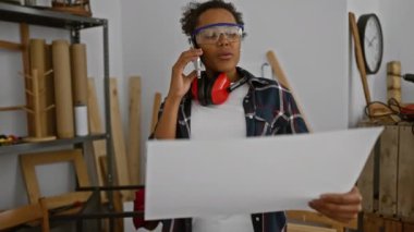 Güvenlik gözlüklü genç bir kadın marangozluk atölyesinde proje planını gözden geçiriyor..