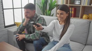 Uzaktan kumandalı bir kadın, gülümseyen bir adam, modern bir dairede, parlak bir oturma odasında, kanepede sıkılmış bir adam..