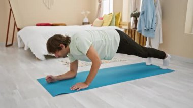 Genç, atletik bir adam mavi bir paspasın üzerinde egzersiz yapıyor. İyi aydınlatılmış bir yatak odasında, aktif bir yaşam tarzı ve ev egzersizi sergiliyor..
