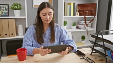 Genç bir İspanyol kadın kapalı ofis ortamında çalışıyor, elinde tablet var ve üzerinde çizgili bir tişört ve odaklanmış bir ifade var..