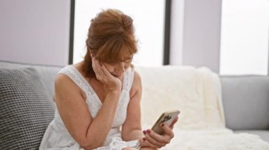 Rahat, modern bir oturma odasında akıllı telefon kullanan olgun, endişeli bir kadın..