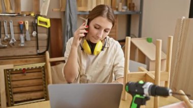 Marangozluk atölyesinde çalışan esmer bir kadın telefon ve dizüstü bilgisayar kullanıyor. Etrafında aletler ve odun var..