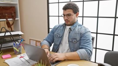 Modern bir ofiste çalışan sakallı genç bir adam. Yanında evrak ve ofis malzemeleri olan bir dizüstü bilgisayar kullanıyor..