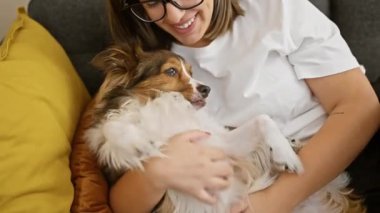 Gözlüklü genç bir kadın kanepede tüylü köpeğiyle sarılıp sıcak ve rahat bir hayat sunuyor..