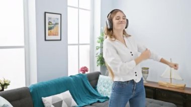 Neşeli genç bir kadın modern oturma odasında dans ediyor, kulaklıklarla müzik dinliyor, şık ev dekorlarıyla çevrili..