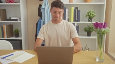 Modern bir ofiste dizüstü bilgisayar kullanan neşeli bir genç adam kitaplar, bardaklar ve çiçeklerle donatılmış..