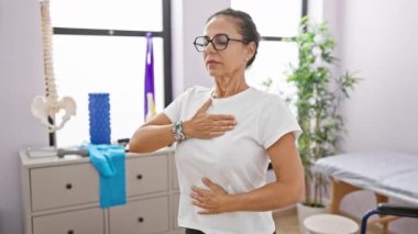 Gözlüklü olgun bir İspanyol kadın tıbbi ekipmanlarla iyi aydınlatılmış bir rehabilitasyon merkezinde nefes egzersizi yapıyor..