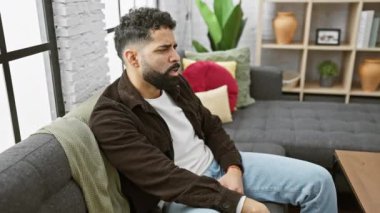 Evde, bıkkınlıktan esneyen yorgun bir genç adam, elleriyle ağzını kapatıyor, uykusuzluk hissi veriyor.