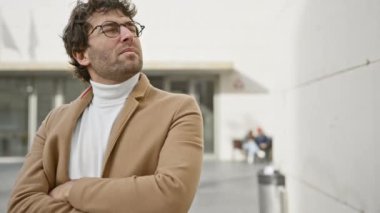 Sakallı, gözlüklü, kollarını kavuşturmuş, şehir ortamında kendine güveni tam İspanyol bir adam..